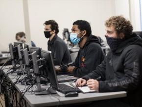 Students sit in a classroom in front of computers while wearing masks